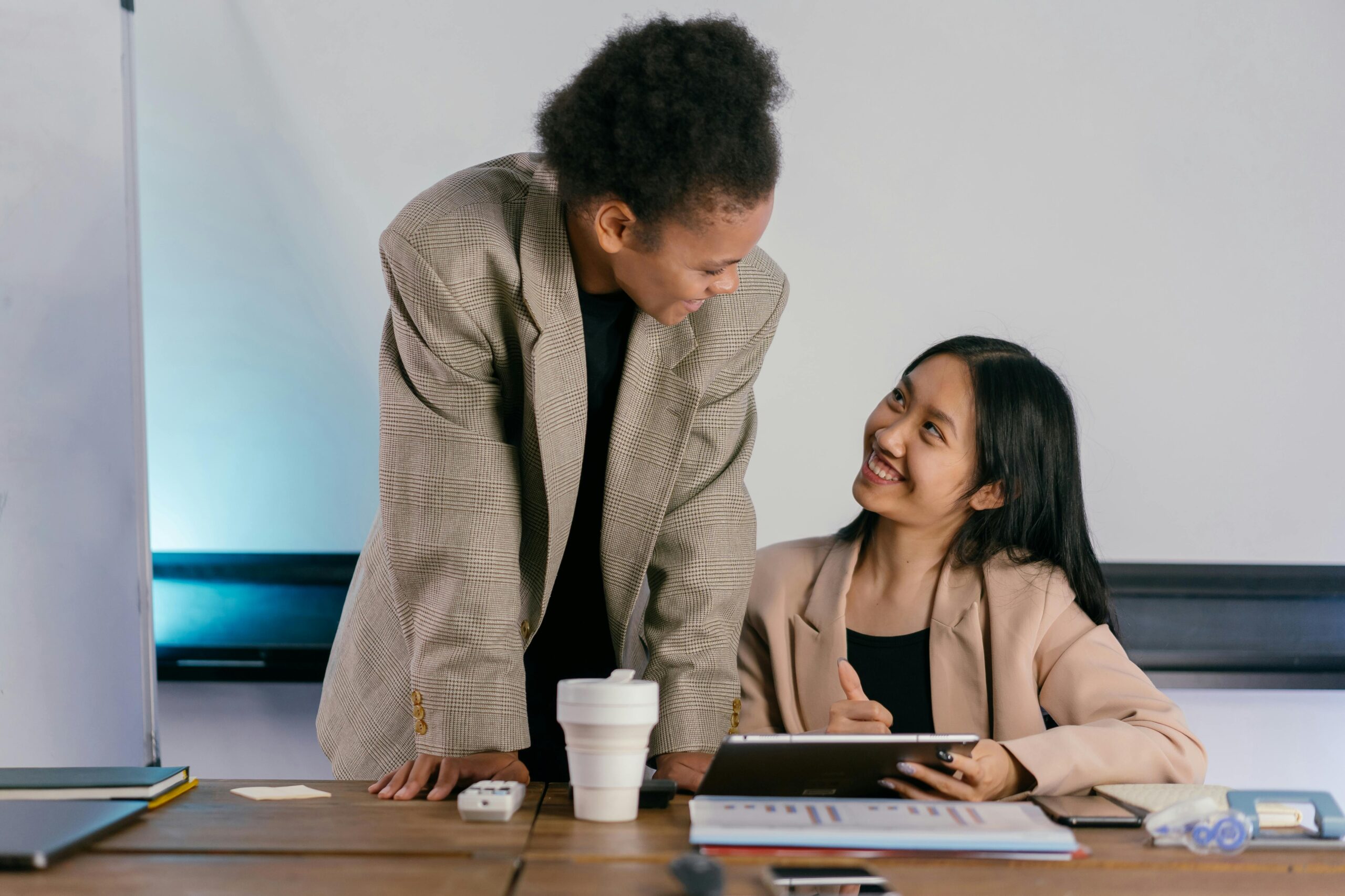 Two colleagues smiling and collaborating at a desk with a tablet and coffee cup.