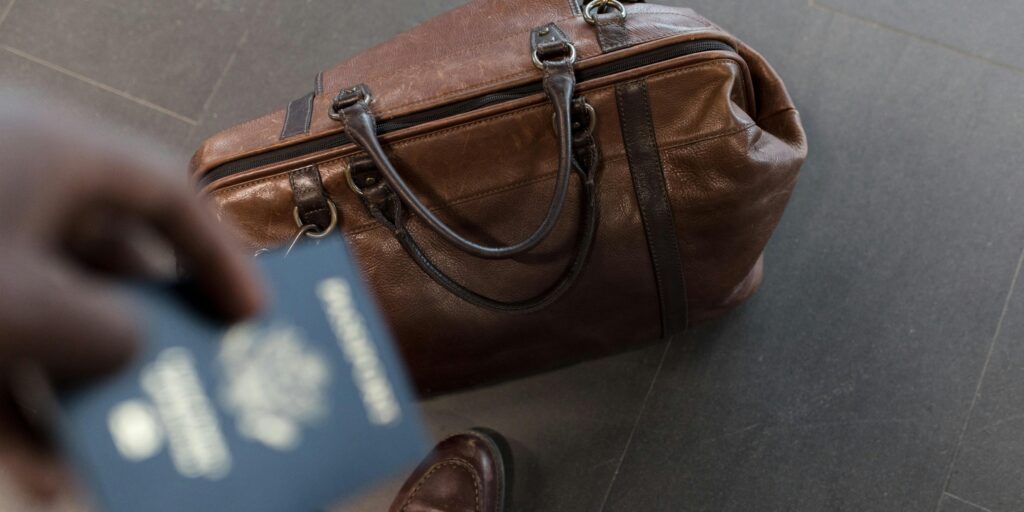 Person holding a passport next to a brown leather travel bag on the floor.